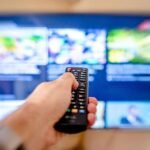Hand holding remote control pointing at a blurred TV screen in a cozy indoor setting.